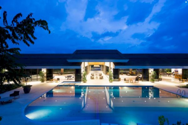 The image shows a luxurious outdoor pool area at dusk, surrounded by modern architecture and lit up under a deep blue sky.