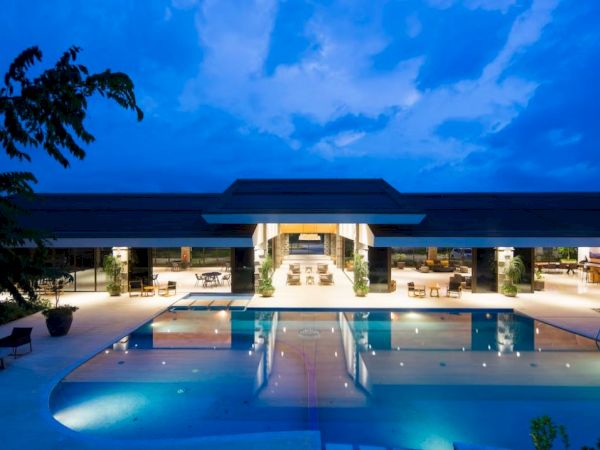 The image shows a luxurious outdoor pool area at dusk, surrounded by modern architecture and lit up under a deep blue sky.