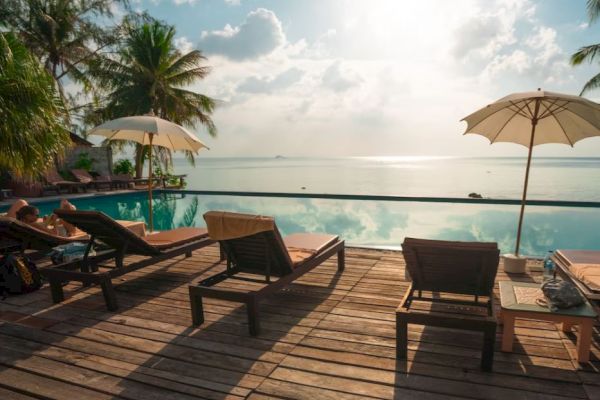 Poolside with sun loungers and umbrellas overlooking the ocean, surrounded by palm trees, under a partly cloudy sky.