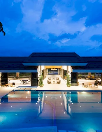 The image shows a luxurious building with a lit swimming pool in front, set against a dark, cloudy evening sky.