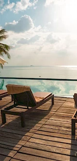 A serene poolside scene with lounge chairs, umbrellas, palm trees, and a view of the ocean under a bright sky.