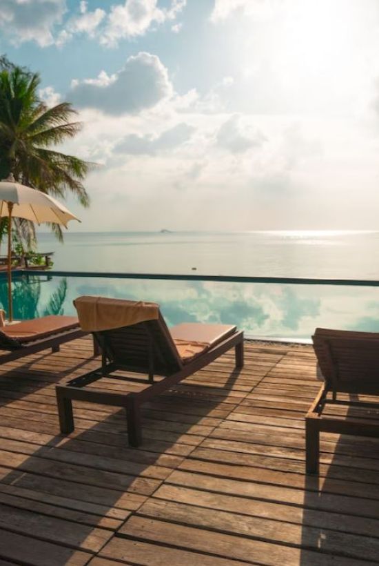 A serene poolside view with lounge chairs, umbrellas, palm trees, and a distant horizon over calm waters.