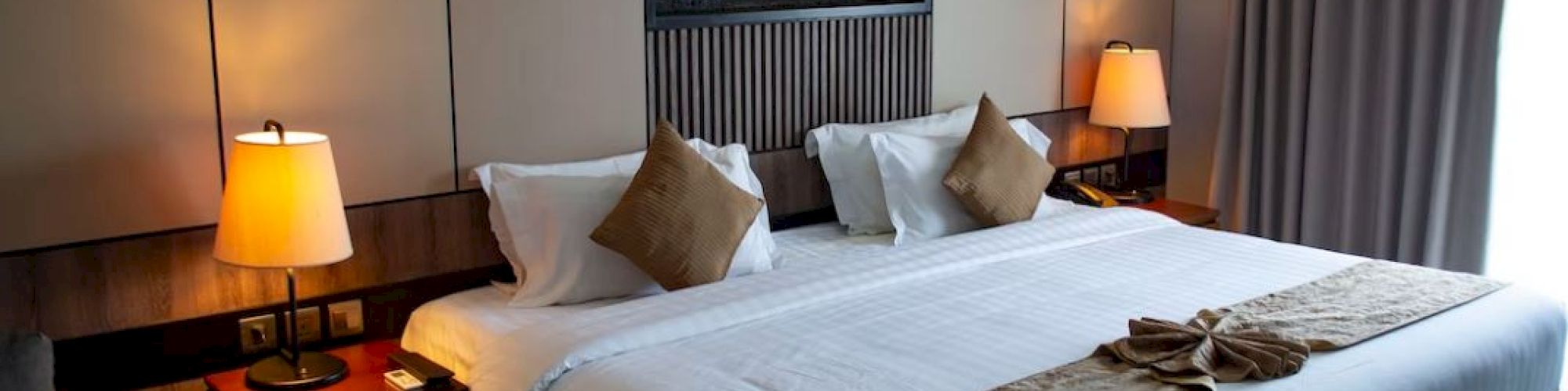 A neatly made hotel room features a large bed with brown accents, two side lamps, a small table, and a window with closed curtains.
