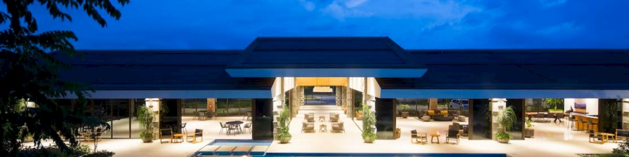 The image shows a luxurious building with an illuminated swimming pool and seating area at dusk, under a cloudy blue sky.