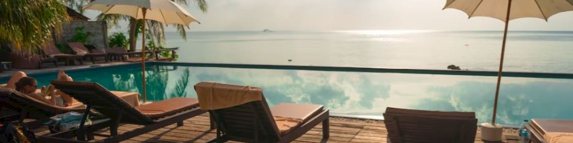 A serene poolside scene with lounge chairs, umbrellas, and palm trees overlooking a calm body of water under a partly cloudy sky.