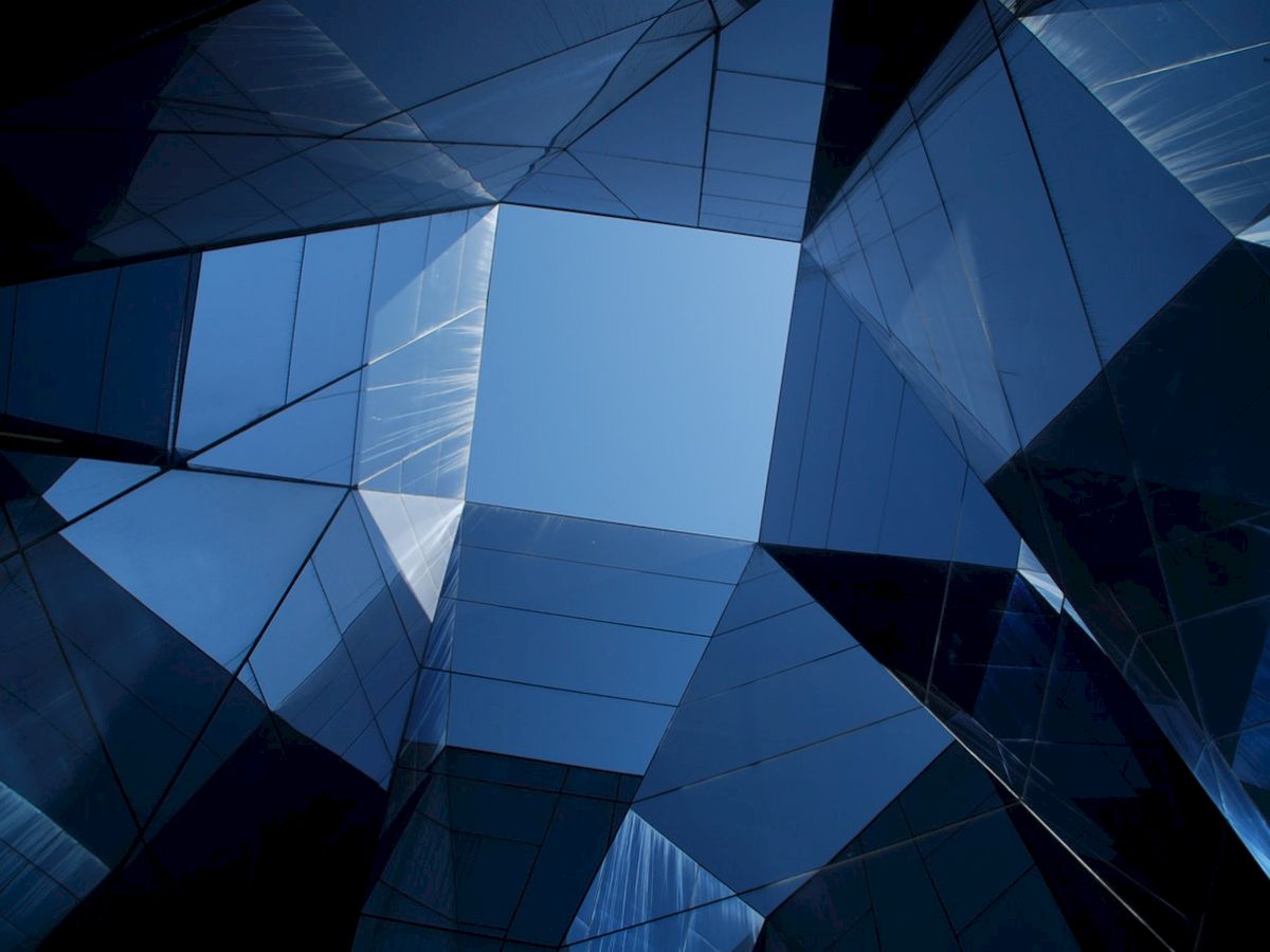 The image shows a view up through reflective, angular glass surfaces with a visible patch of blue sky in the center.