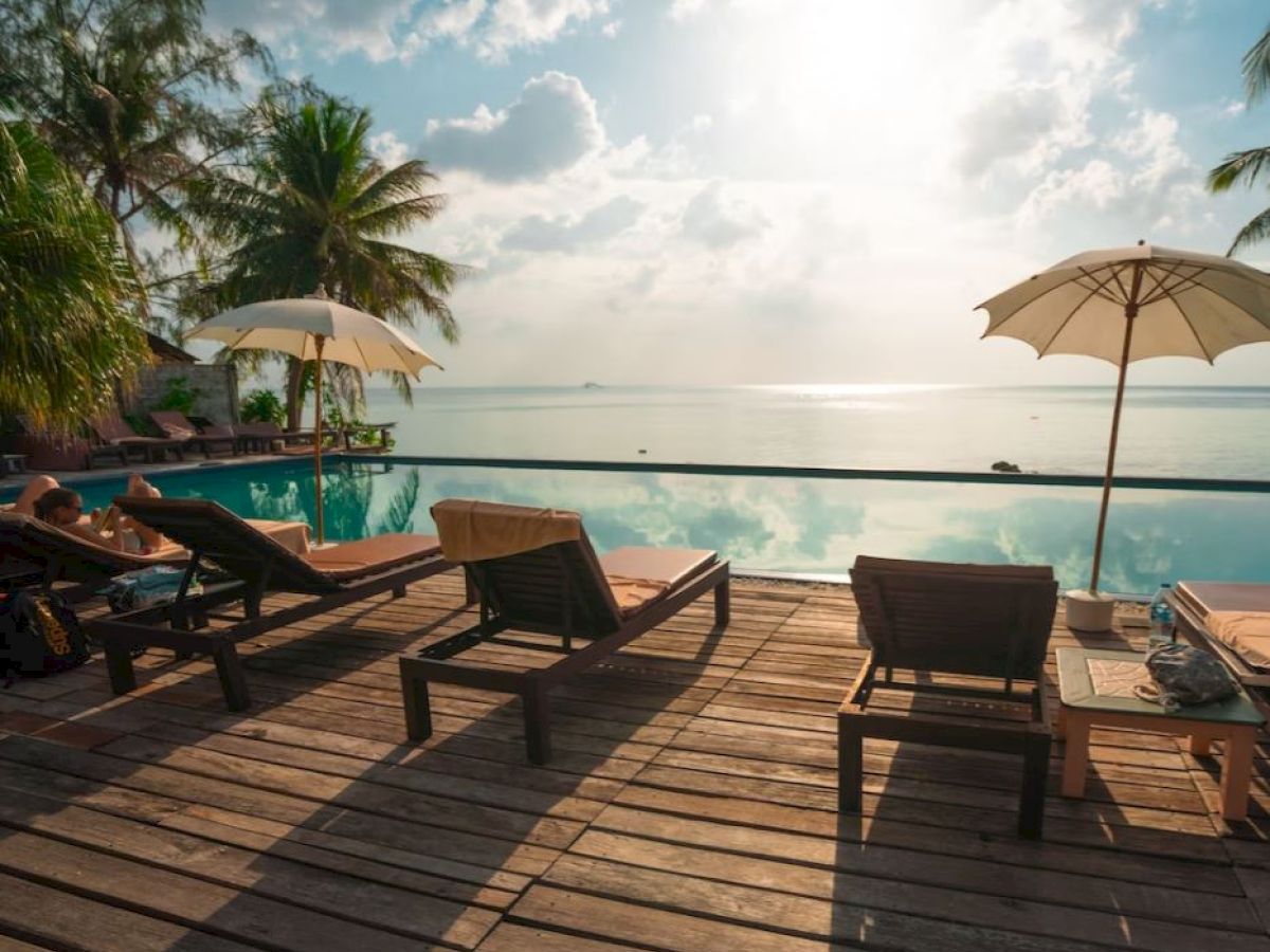 A serene poolside view with lounge chairs, parasols, and palm trees overlooking a calm body of water under a partly cloudy sky.