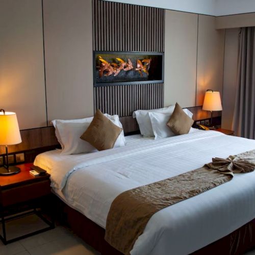 A cozy hotel room with a large bed, decorative pillows, lit lamps, and a wall-mounted art piece above.