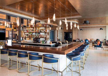 A modern bar with a wooden counter, blue stools, hanging lights, and a well-stocked liquor shelf in a spacious, stylish setting.
