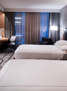The image shows a modern hotel room with two beds, a flat-screen TV, a desk, and patterned wallpaper, creating a comfortable, stylish atmosphere.