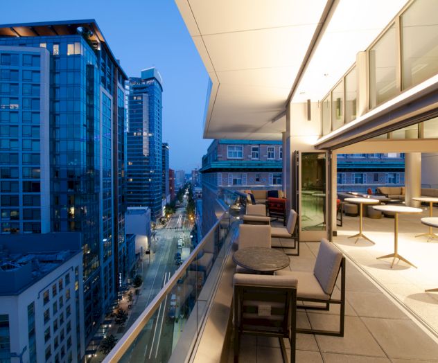 A modern rooftop terrace with seating overlooks a cityscape of tall buildings at dusk, with lights from the buildings illuminating the scene.