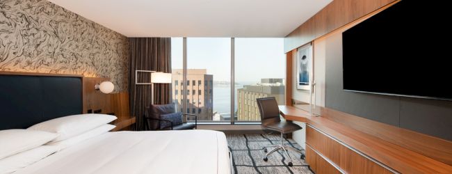 A modern hotel room with a large bed, desk, chair, and TV, featuring floor-to-ceiling windows with a cityscape view.