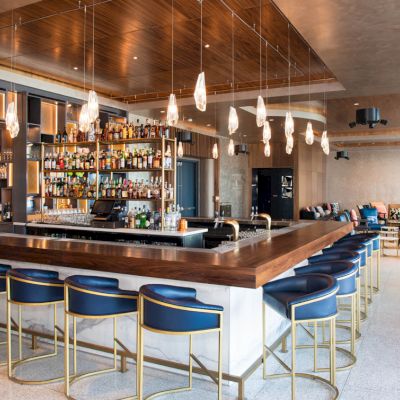 This image shows a modern bar with a central island, blue bar stools, and a variety of bottles. Pendant lights hang from the ceiling.
