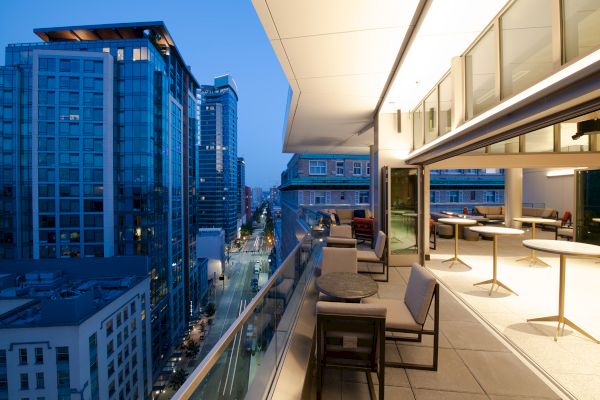 A modern urban balcony overlooks a city street below, featuring outdoor seating and large windows, all illuminated by evening lights.