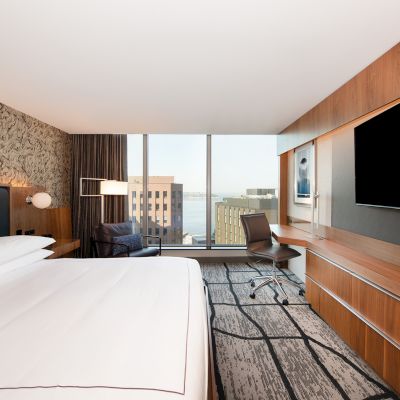 A modern hotel room with a large bed, TV, desk, and city view through a window, featuring contemporary decor and design elements.