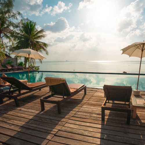 Sunlit deck with lounge chairs and umbrellas by an infinity pool, overlooking a serene ocean and scattered clouds.