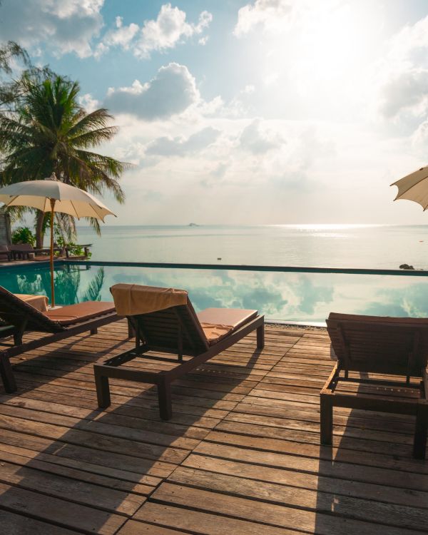 Sunlit deck with lounge chairs and umbrellas by an infinity pool, overlooking a serene ocean and scattered clouds.