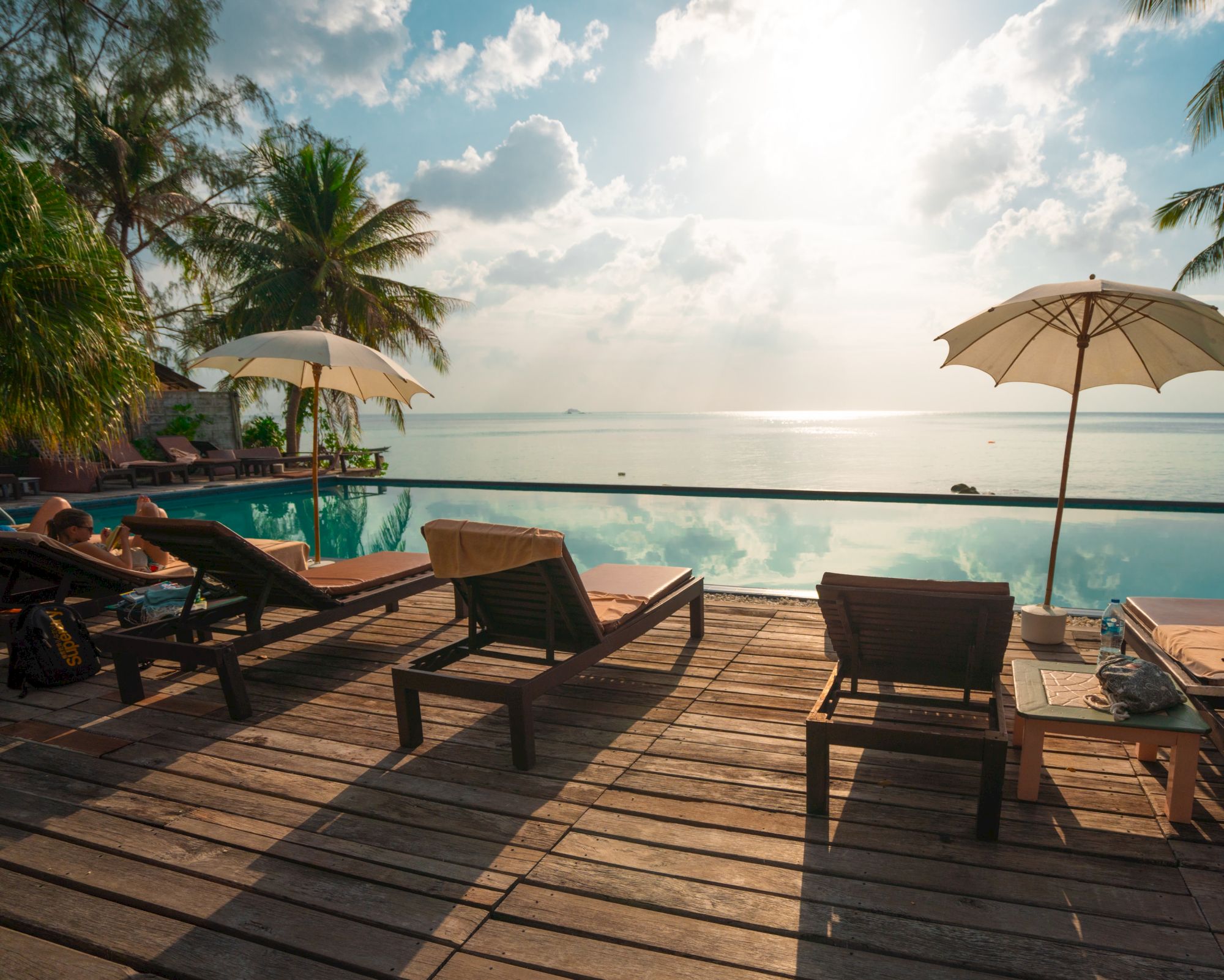 Wooden deck with lounge chairs, umbrellas overlooking an infinity pool and serene ocean view under a partly cloudy sky.