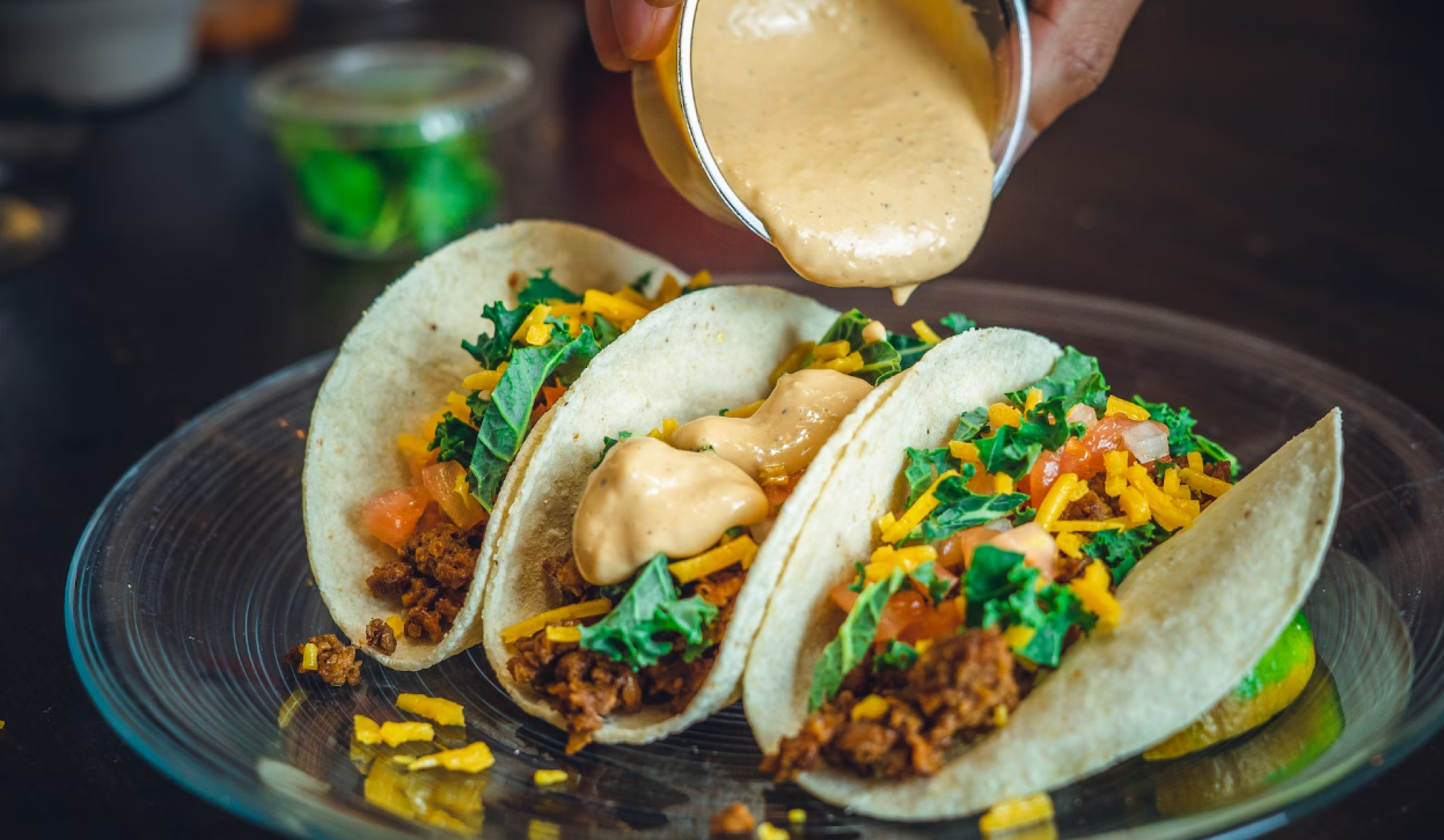 Three tacos filled with greens, cheese, and meat are being topped with a creamy sauce on a clear plate.