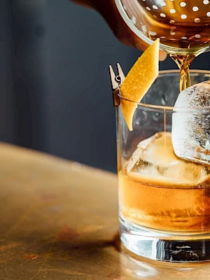 A cocktail being poured over ice in a glass with a citrus twist garnish on a bar counter.