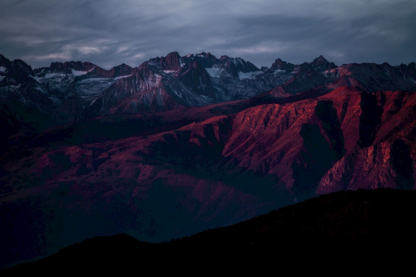 A mountain range is bathed in red sunlight with a cloudy sky above, creating a dramatic and scenic landscape.