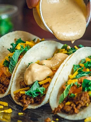 Three tacos filled with ground meat, lettuce, tomatoes, and cheese, being topped with a creamy sauce on a plate.