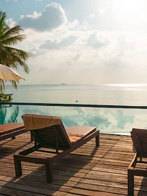 Poolside loungers and umbrellas overlook a tranquil sea view, surrounded by palm trees, under a partly cloudy sky.
