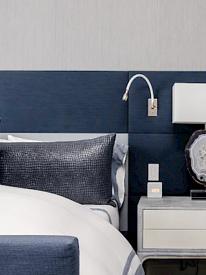 The image features a modern bedroom with a blue and gray color scheme, a bed with pillows, and a nightstand with a lamp and decor piece.