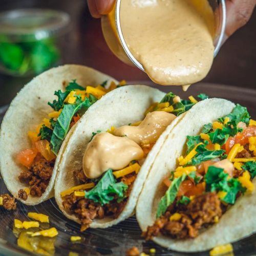 Three tacos filled with ground meat, shredded cheese, kale, and diced tomatoes, topped with creamy sauce being poured over them.