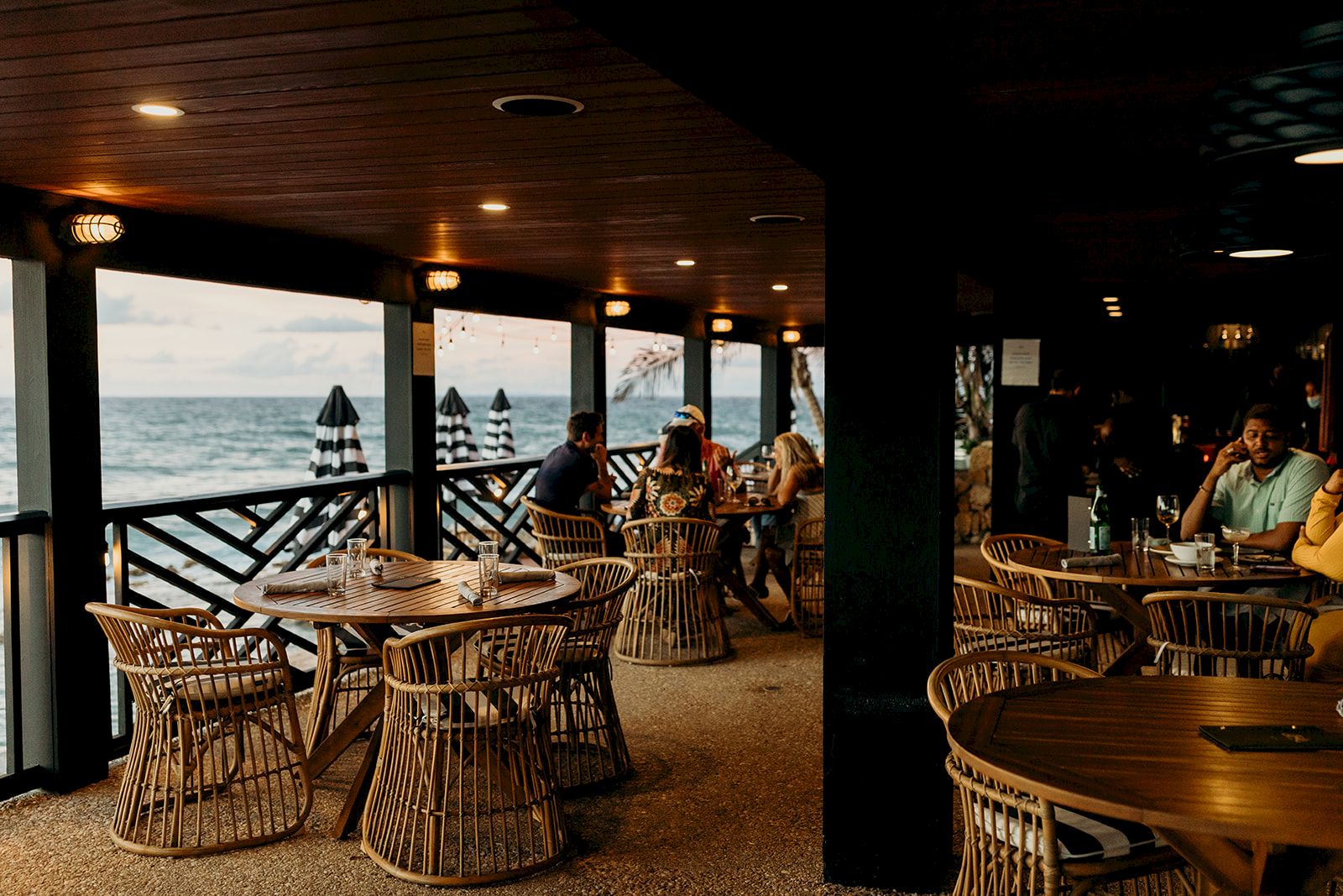An outdoor restaurant with wicker chairs overlooks the ocean. People are seated, enjoying the view as sunlight filters through the canopy.