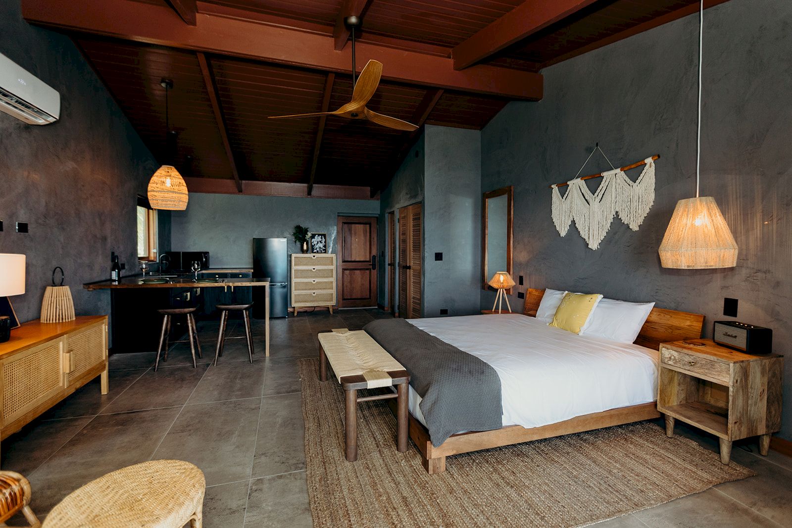 A cozy bedroom with a double bed, wooden furniture, a small kitchen area, pendant lighting, and a rustic decor theme.