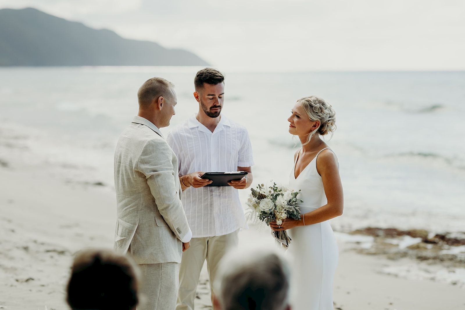 A couple is getting married on a beach, with an officiant and guests present, against a backdrop of the ocean and mountains.