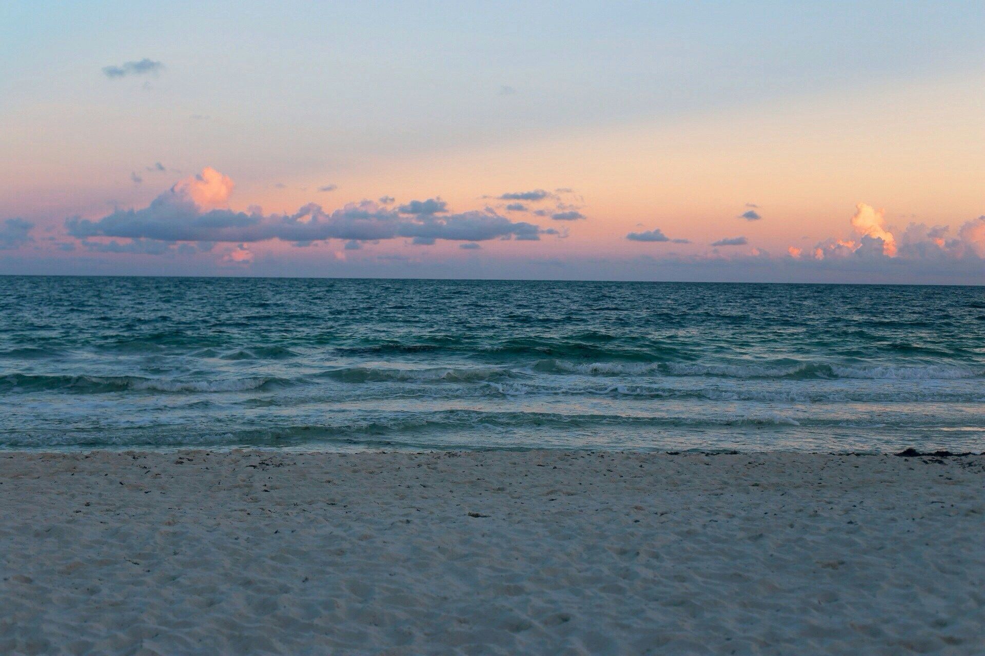 A serene beach scene with gentle waves under a colorful, cloudy sunset sky. The sandy shore is in the foreground.