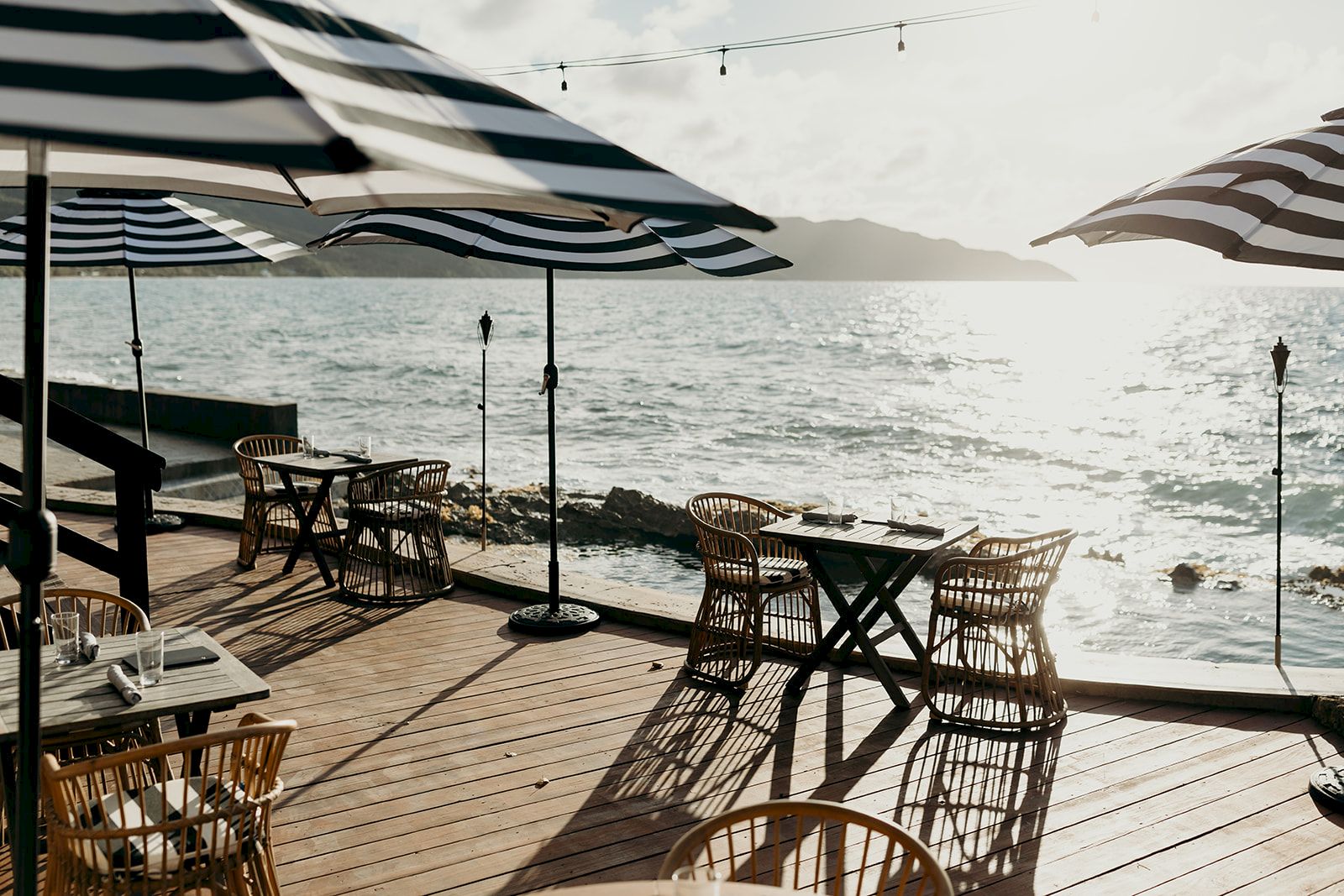 A seaside deck with tables, chairs, and striped umbrellas overlooks the ocean at sunset, creating a serene and inviting atmosphere.