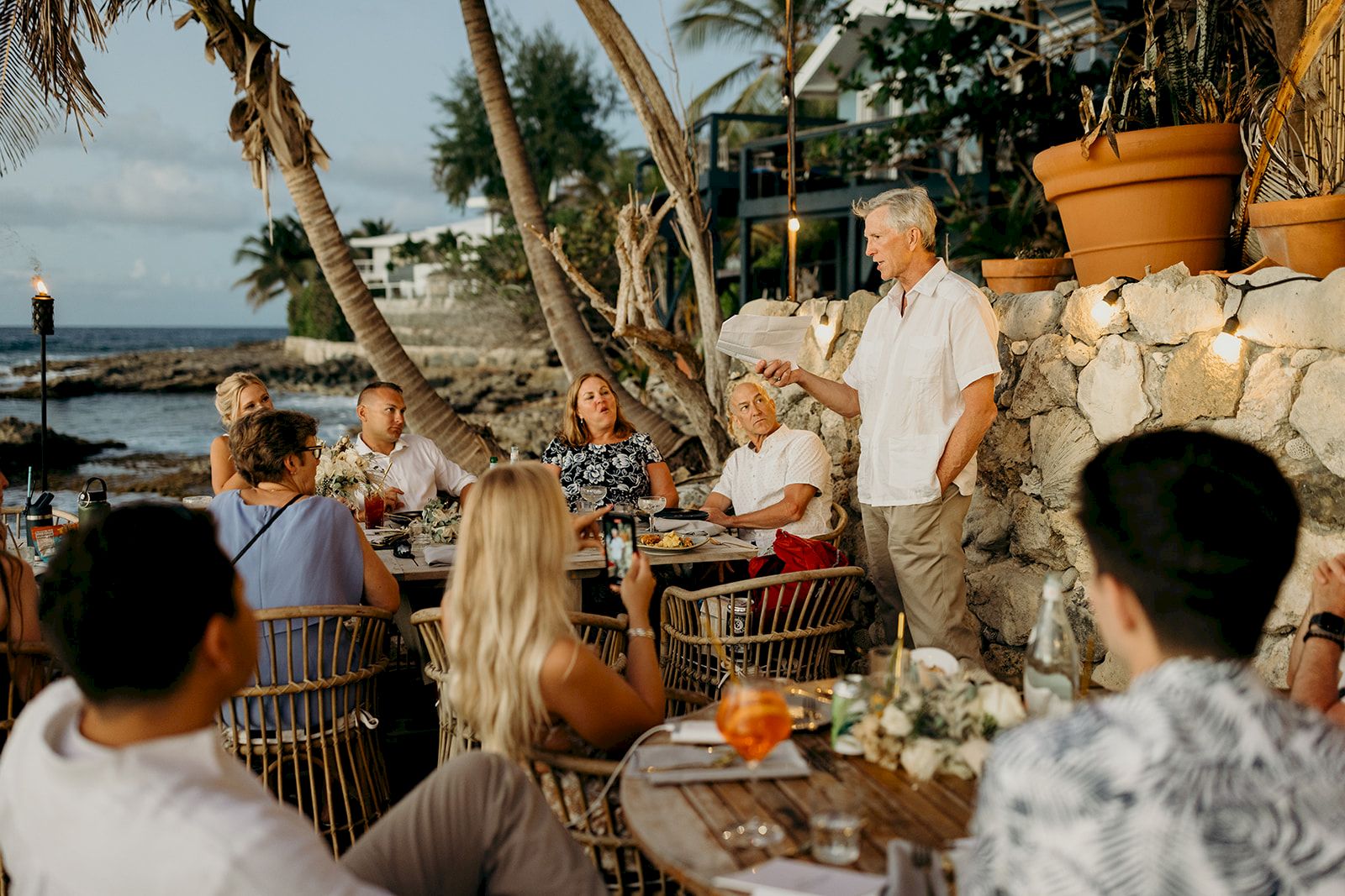 A group of people dine outdoors near the ocean, with a man standing and speaking, surrounded by palm trees and a stone wall.