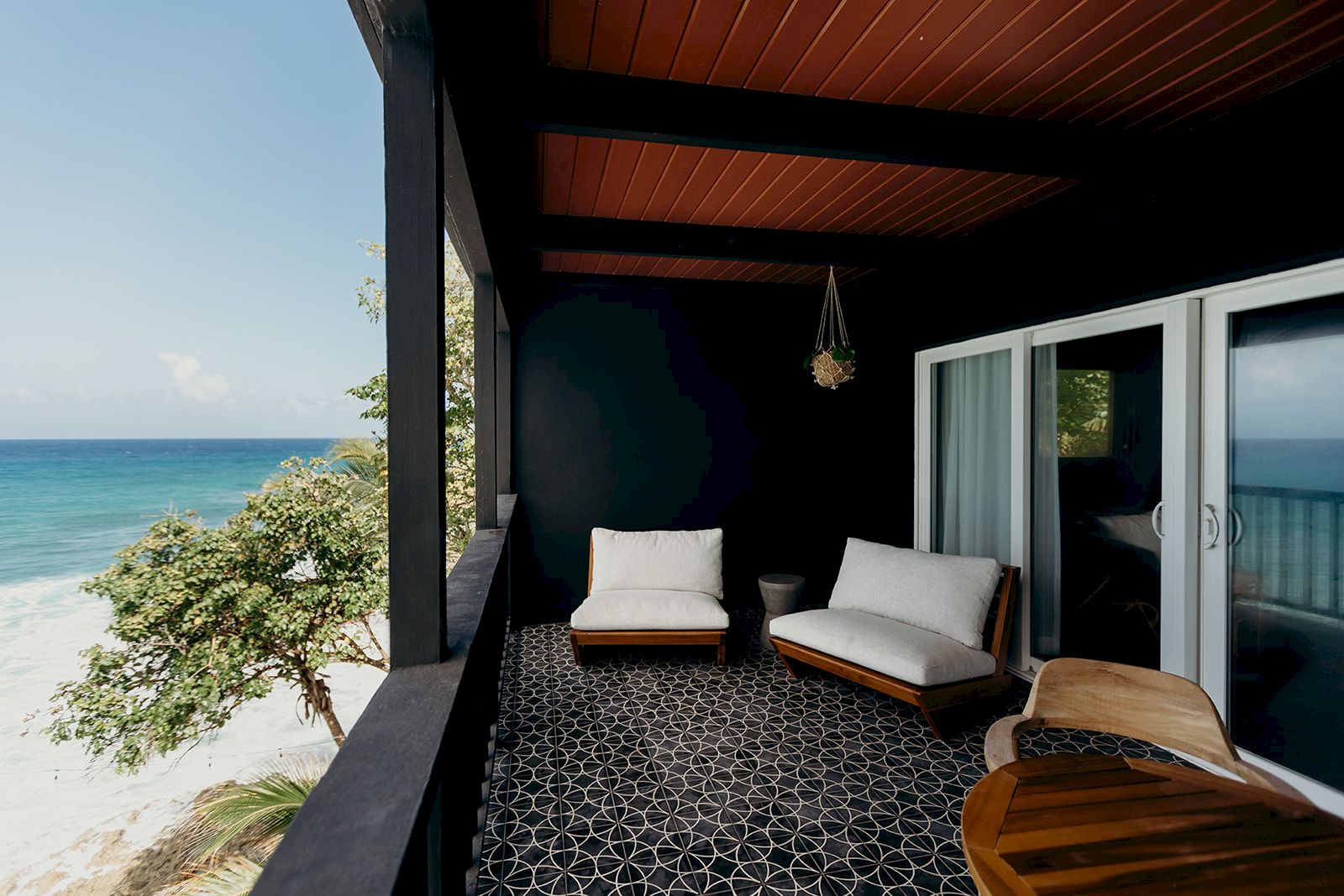 A beachside balcony with chairs, a geometric tile floor, and ocean view, enclosed by dark walls and a wooden ceiling, under a clear sky.