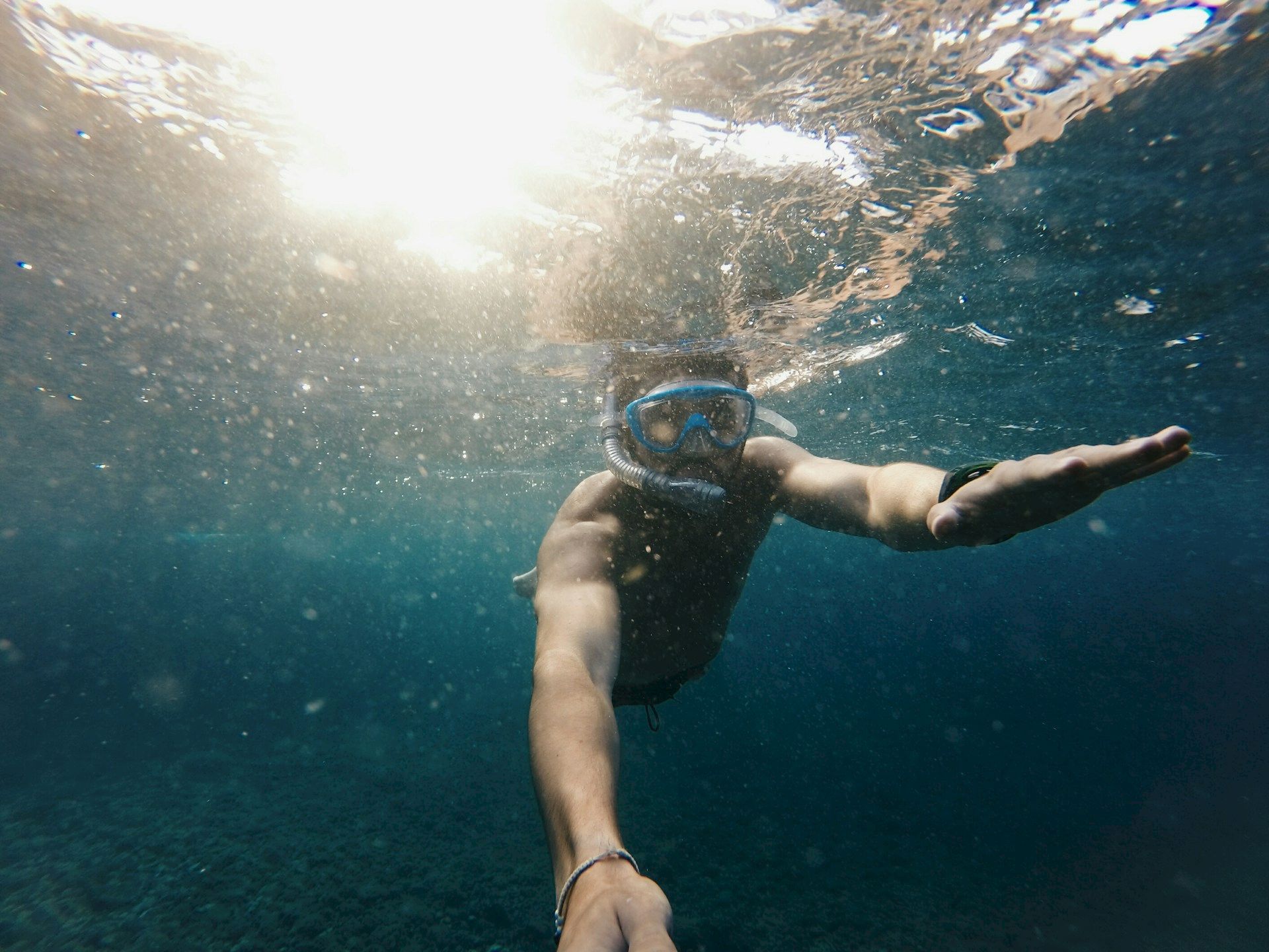 A person is underwater, wearing a snorkel mask, swimming with a sunlight reflection above, capturing the moment with an extended arm.