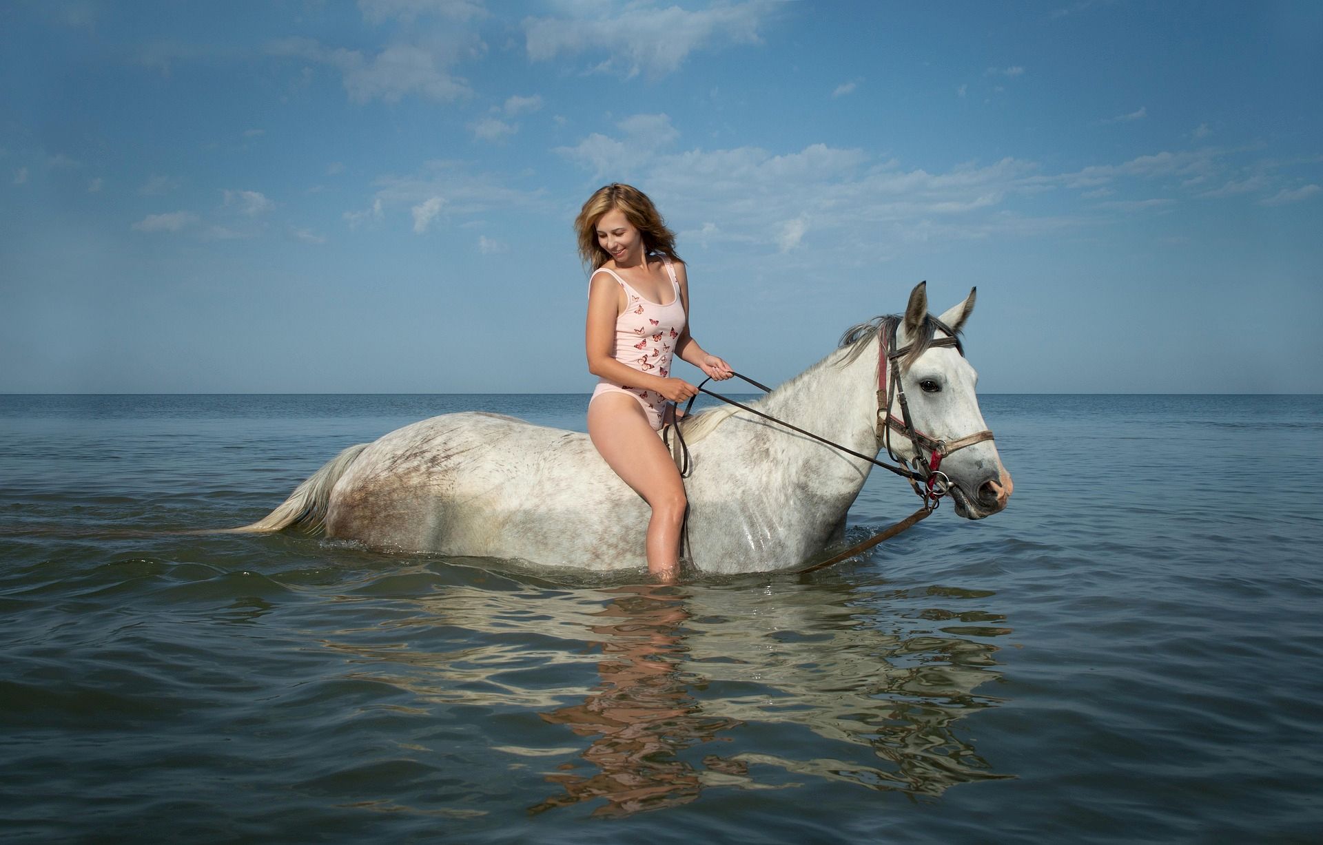 A person in swimwear is riding a white horse in the sea, with a clear sky and calm water.