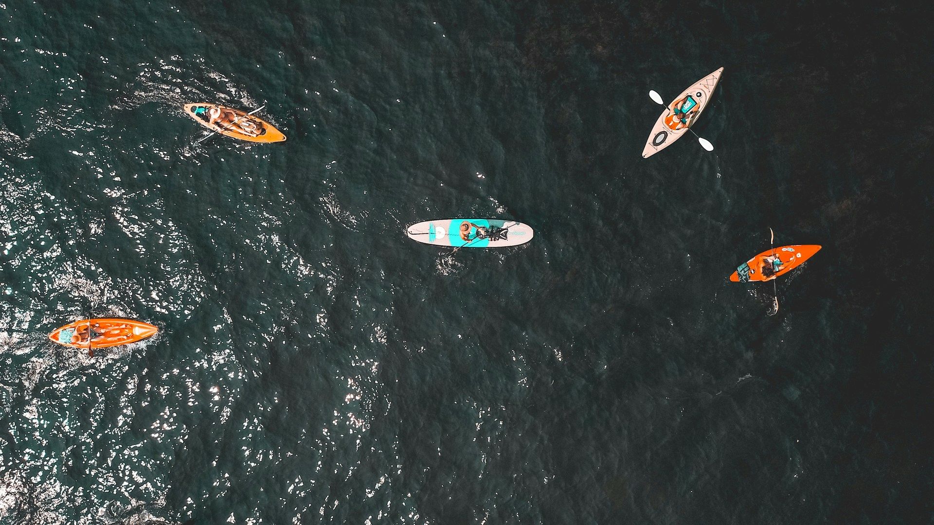 The image shows five people kayaking on open water, captured from an aerial view, showcasing various colorful kayaks.