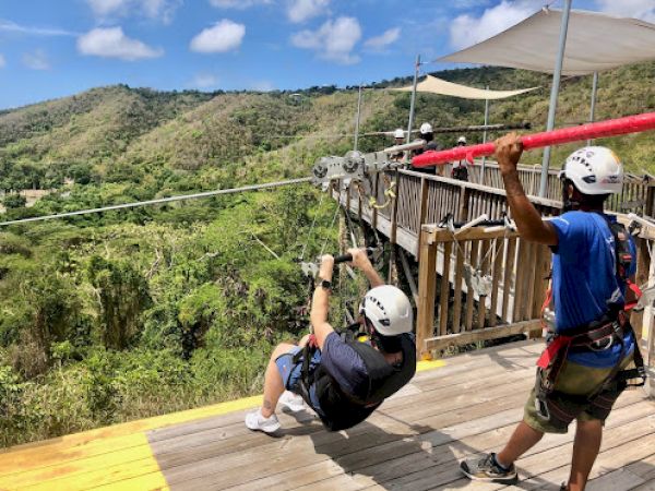 Two people are at a zipline platform. One is seated, ready to launch, while the other assists. Bright day with lush hills in the background.