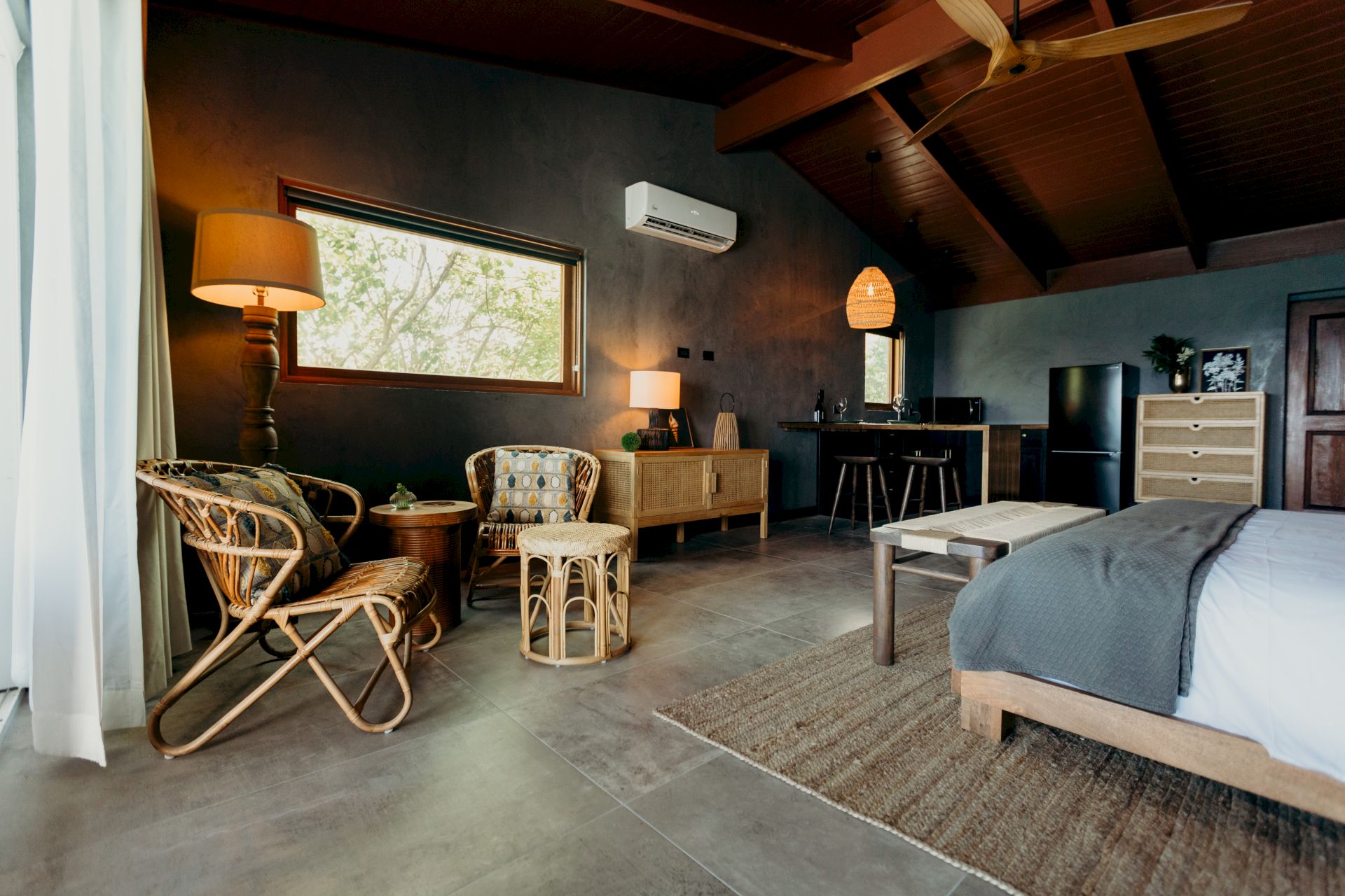 A cozy bedroom with wicker chairs, bed, rug, and a small kitchenette, featuring wooden ceiling beams and soft lighting.
