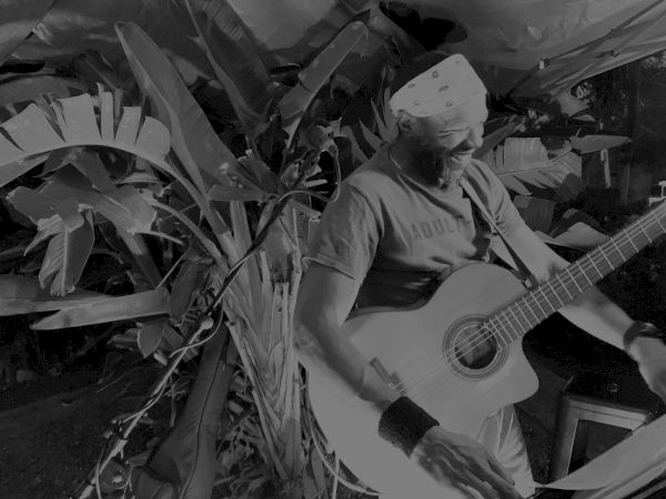 A person is playing a guitar outdoors, surrounded by large leafy plants and wearing a bandana and armbands.