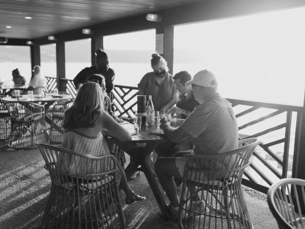 People sitting at tables in an outdoor dining area with sunlight streaming in, engaged in conversation and enjoying the view.