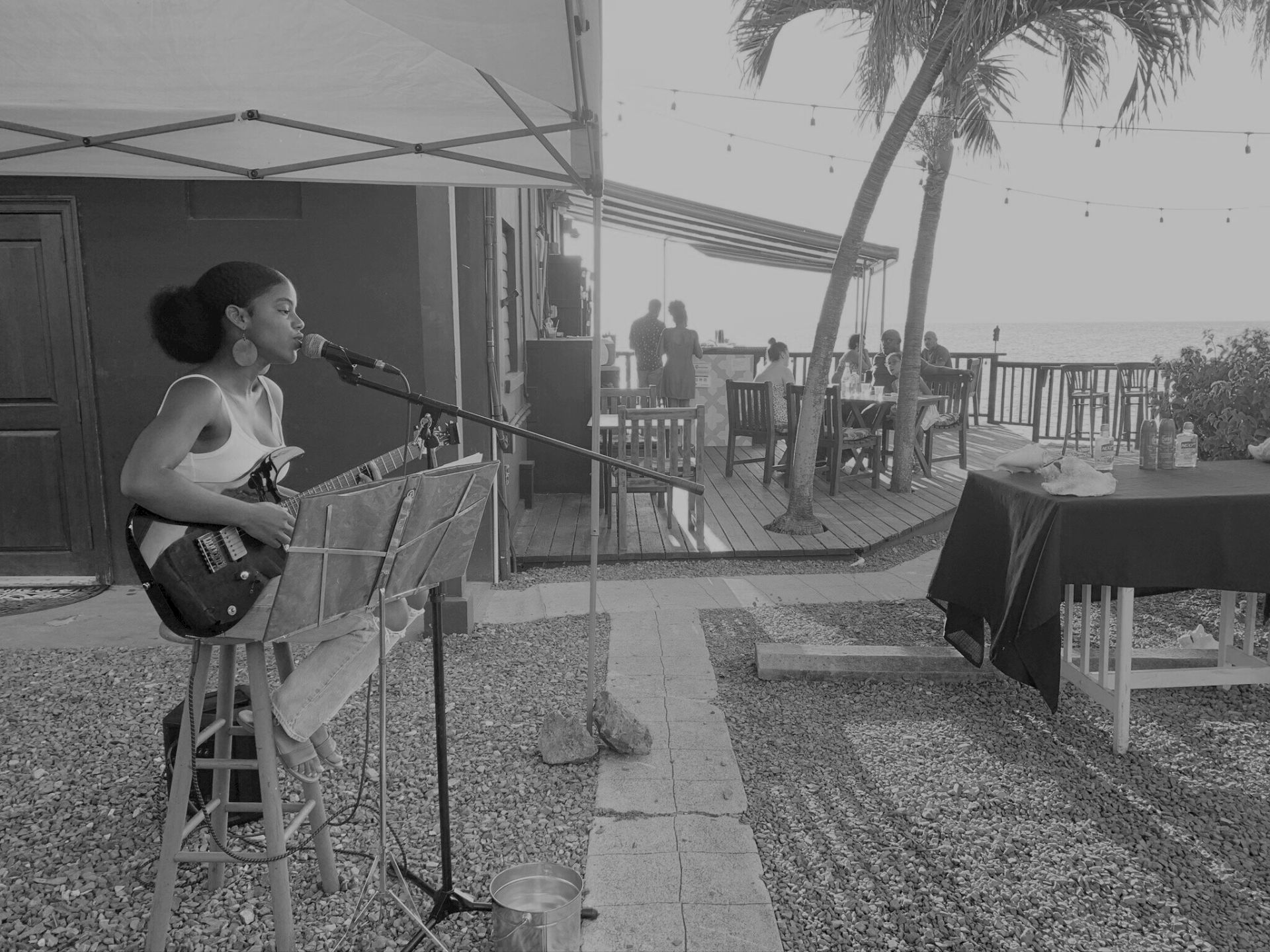 A person is playing guitar and singing into a microphone outdoors, with tables and people in the background, near the ocean.