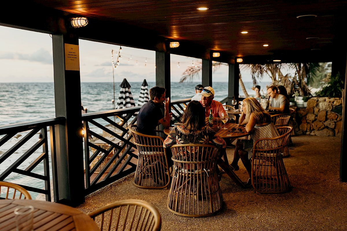A group of people are sitting around a table on a covered patio by the sea, enjoying drinks and conversation as the sun sets.