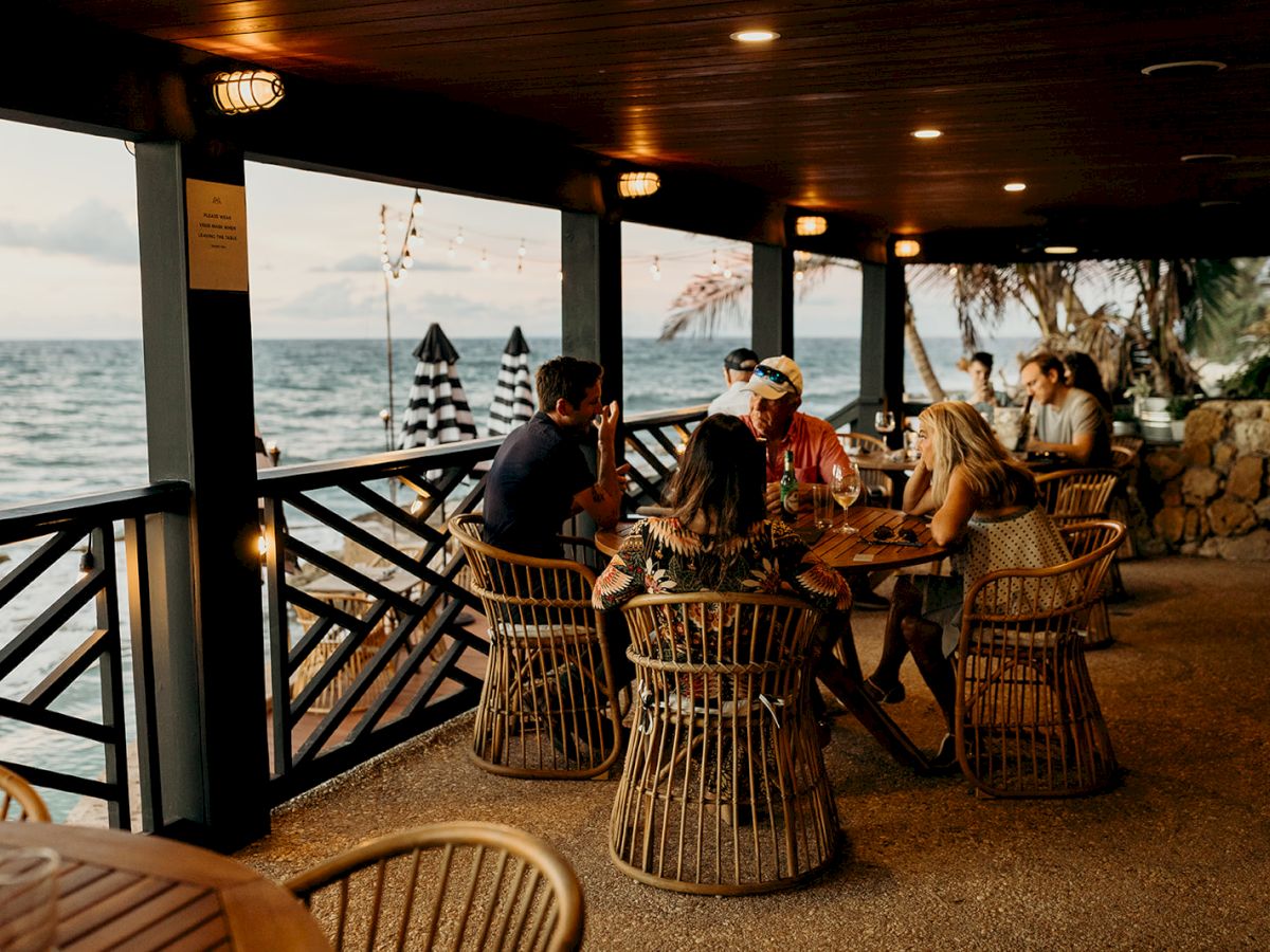 A group of people are sitting around a table on a covered patio by the sea, enjoying drinks and conversation as the sun sets.
