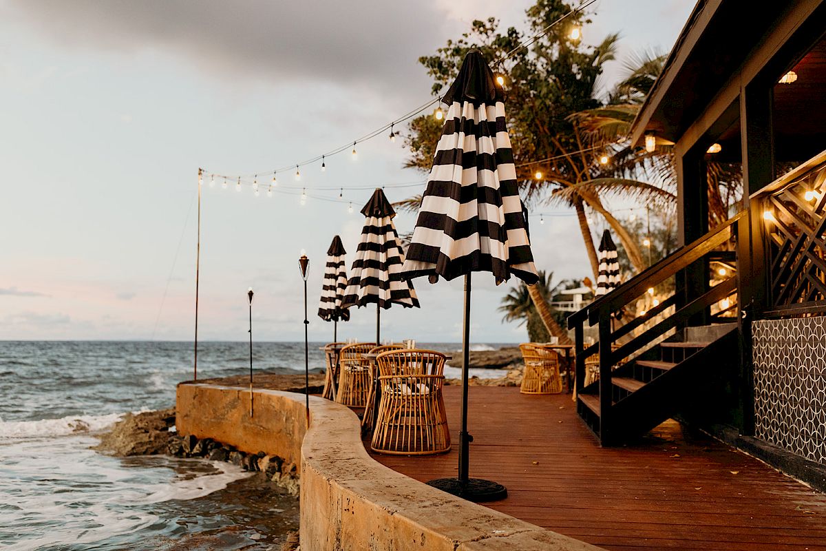 A beachfront deck with black-and-white striped umbrellas, wicker chairs, string lights, and palm trees overlooking the ocean.