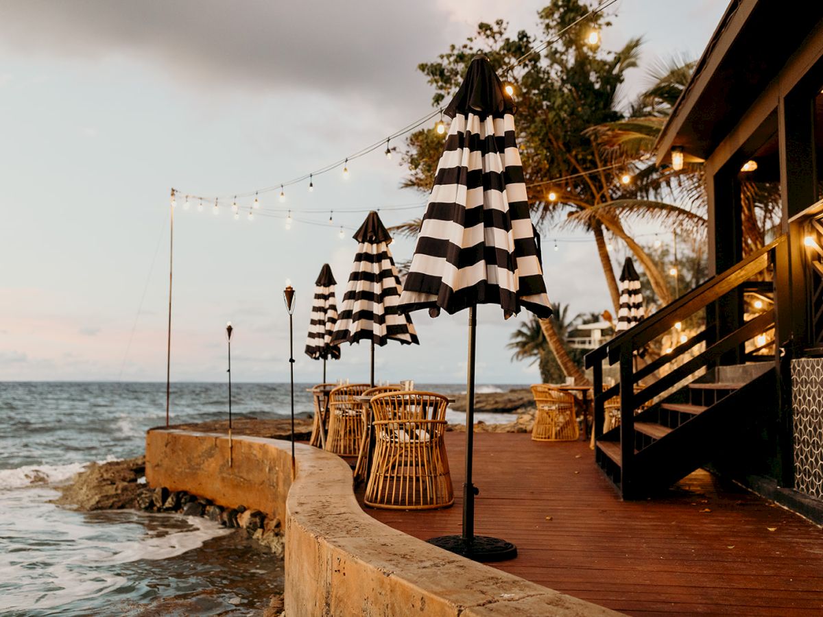 A beachfront deck with black-and-white striped umbrellas, wicker chairs, string lights, and palm trees overlooking the ocean.