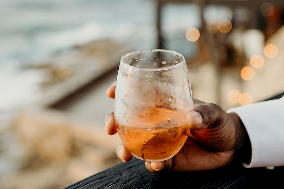 A hand holds a glass of amber liquid, possibly a drink, with a blurred scenic background, suggesting a relaxed outdoor setting.