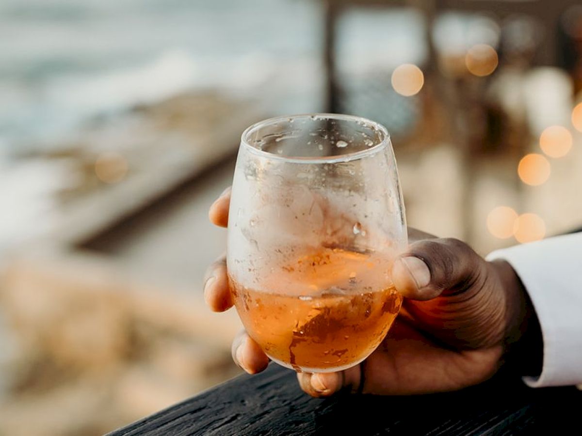 A hand holds a glass of amber liquid, possibly a drink, with a blurred scenic background, suggesting a relaxed outdoor setting.